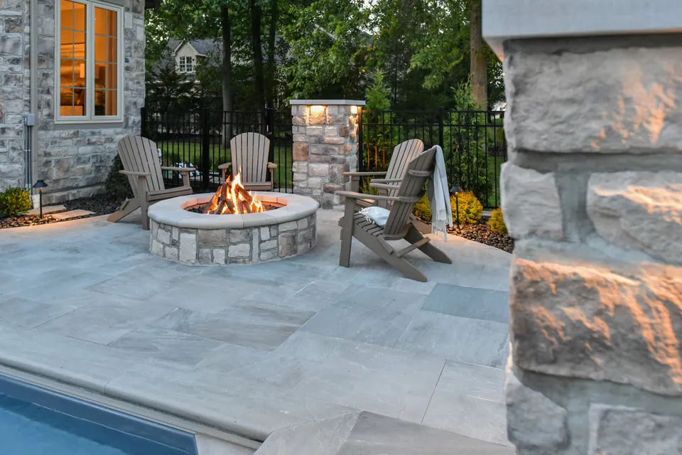 Poolside firepit seating framed by stone piers, paver deck, house wall, and planted edge